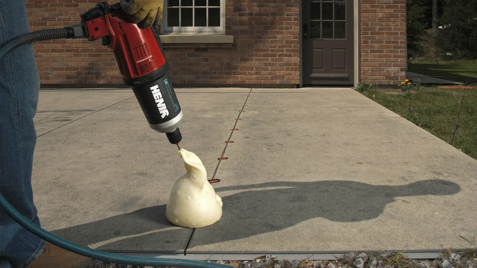 Technician injecting polyurethane foam through small drilled ports to lift a settled concrete driveway section back level outside an Oklahoma home